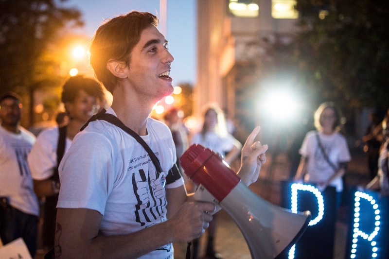 A white man holding a megaphone, centered, stands among a group of students at night.