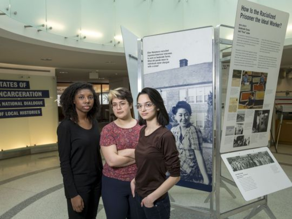 Three women standing in front of the States of Incarceration exhibit in the lobby of a public university library